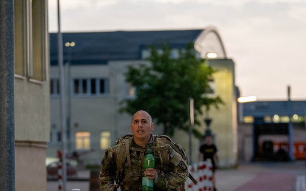 A bald male soldier in MultiCam/OCP camouflage uniform is running or fast-walking along a paved pathway on what appears to be a military installation. He is carrying a tactical backpack/rucksack and holding a green plastic water bottle in his right hand. The setting appears to be dusk or early morning, with institutional buildings and red-and-white bollards lining the path in the background.
