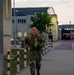 A bald male soldier in MultiCam/OCP camouflage uniform is running or fast-walking along a paved pathway on what appears to be a military installation. He is carrying a tactical backpack/rucksack and holding a green plastic water bottle in his right hand. The setting appears to be dusk or early morning, with institutional buildings and red-and-white bollards lining the path in the background.