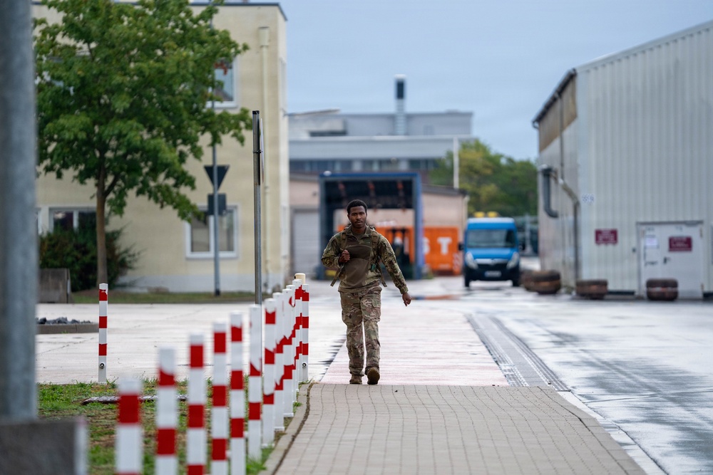 A soldier in camouflage military uniform is jogging along a paved walkway on what appears to be a military installation or industrial compound. The area features red-and-white bollards lining the path, institutional buildings, and a blue van visible in the background. The overcast weather and wet surfaces suggest recent rainfall.