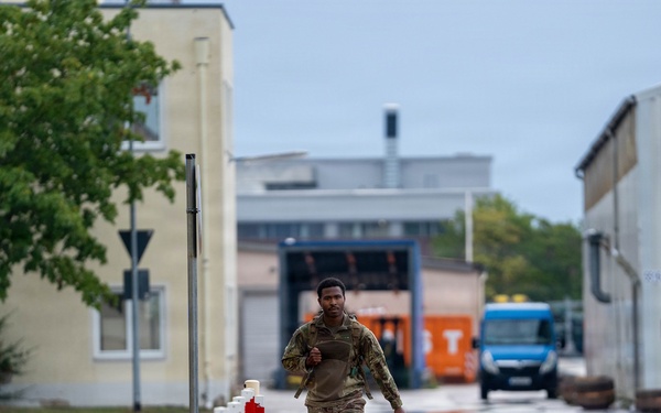 A soldier in camouflage military uniform is jogging along a paved walkway on what appears to be a military installation or industrial compound. The area features red-and-white bollards lining the path, institutional buildings, and a blue van visible in the background. The overcast weather and wet surfaces suggest recent rainfall.