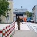 A soldier in camouflage military uniform is jogging along a paved walkway on what appears to be a military installation or industrial compound. The area features red-and-white bollards lining the path, institutional buildings, and a blue van visible in the background. The overcast weather and wet surfaces suggest recent rainfall.