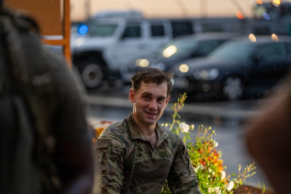 A young male soldier wearing US Army OCP (Operational Camouflage Pattern) uniform is smiling while engaged in conversation with others. The scene appears to be outdoors during dusk or dawn in light rain, with a parking area containing military or government vehicles visible in the blurred background. The atmosphere appears relaxed and informal.