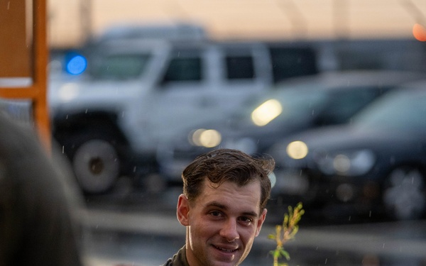 A young male soldier wearing US Army OCP (Operational Camouflage Pattern) uniform is smiling while engaged in conversation with others. The scene appears to be outdoors during dusk or dawn in light rain, with a parking area containing military or government vehicles visible in the blurred background. The atmosphere appears relaxed and informal.