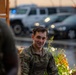 A young male soldier wearing US Army OCP (Operational Camouflage Pattern) uniform is smiling while engaged in conversation with others. The scene appears to be outdoors during dusk or dawn in light rain, with a parking area containing military or government vehicles visible in the blurred background. The atmosphere appears relaxed and informal.