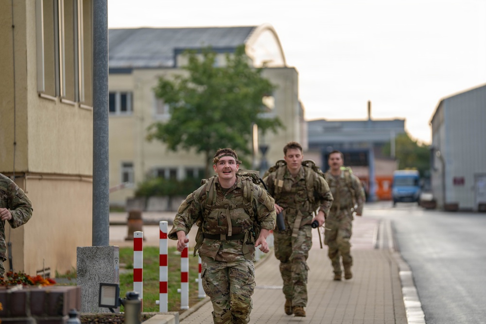 Several military personnel in camouflage uniforms are running or fast-marching along a paved pathway on what appears to be a military installation or base. The soldiers are carrying large rucksacks on their backs, suggesting a ruck march or physical training exercise. The setting includes institutional buildings and infrastructure consistent with a European military base.