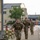 12th Combat Aviation Brigade hosts Danish Flyver March Several military personnel in camouflage uniforms are running or fast-marching along a paved pathway on what appears to be a military installation or base. The soldiers are carrying large rucksacks on their backs, suggesting a ruck march or physical training exercise. The setting includes institutional buildings and infrastructure consistent with a European military base.