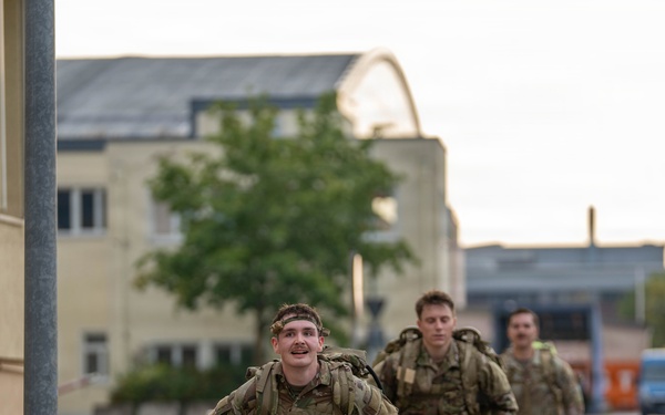 Several military personnel in camouflage uniforms are running or fast-marching along a paved pathway on what appears to be a military installation or base. The soldiers are carrying large rucksacks on their backs, suggesting a ruck march or physical training exercise. The setting includes institutional buildings and infrastructure consistent with a European military base.