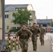 Several military personnel in camouflage uniforms are running or fast-marching along a paved pathway on what appears to be a military installation or base. The soldiers are carrying large rucksacks on their backs, suggesting a ruck march or physical training exercise. The setting includes institutional buildings and infrastructure consistent with a European military base.