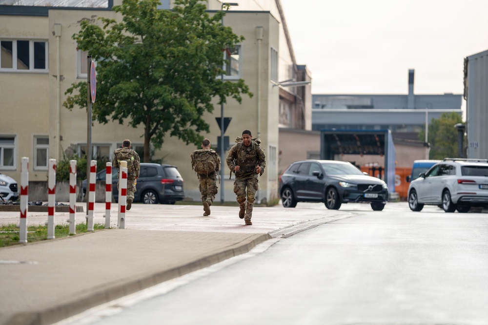 Three military personnel in camouflage uniforms and tactical gear are running along a paved path in what appears to be a military installation or base. The soldiers are carrying backpacks and equipment, suggesting a physical training exercise or ruck march. The setting includes institutional buildings, parked civilian and military vehicles, and European-style road markings.
