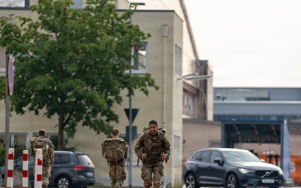 Three military personnel in camouflage uniforms and tactical gear are running along a paved path in what appears to be a military installation or base. The soldiers are carrying backpacks and equipment, suggesting a physical training exercise or ruck march. The setting includes institutional buildings, parked civilian and military vehicles, and European-style road markings.