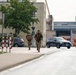 Three military personnel in camouflage uniforms and tactical gear are running along a paved path in what appears to be a military installation or base. The soldiers are carrying backpacks and equipment, suggesting a physical training exercise or ruck march. The setting includes institutional buildings, parked civilian and military vehicles, and European-style road markings.