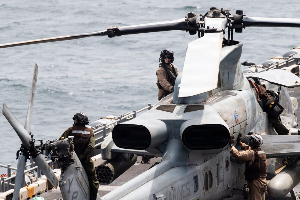 Military personnel in flight gear and helmets are conducting maintenance or pre-flight checks on what appears to be a AH-1Z Viper attack helicopter aboard a naval vessel at sea. The word 'MARINES' is partially visible on the aircraft fuselage, indicating U.S. Marine Corps ownership. At least three crew members are visible working around the helicopter on what appears to be an amphibious ship flight deck.