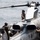 Military personnel in flight gear and helmets are conducting maintenance or pre-flight checks on what appears to be a AH-1Z Viper attack helicopter aboard a naval vessel at sea. The word 'MARINES' is partially visible on the aircraft fuselage, indicating U.S. Marine Corps ownership. At least three crew members are visible working around the helicopter on what appears to be an amphibious ship flight deck.