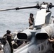 Military personnel in flight gear and helmets are conducting maintenance or pre-flight checks on what appears to be a AH-1Z Viper attack helicopter aboard a naval vessel at sea. The word 'MARINES' is partially visible on the aircraft fuselage, indicating U.S. Marine Corps ownership. At least three crew members are visible working around the helicopter on what appears to be an amphibious ship flight deck.