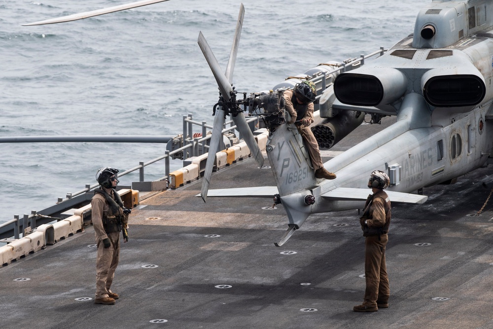 Three military personnel in flight suits and helmets are conducting maintenance or pre-flight checks on a U.S. Marines helicopter on the flight deck of a naval vessel. One crew member is seated on the tail section of the helicopter, while two others stand on the deck observing. The aircraft bears the 'MARINES' marking and bureau number 169251, and the scene takes place at sea.