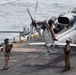Three military personnel in flight suits and helmets are conducting maintenance or pre-flight checks on a U.S. Marines helicopter on the flight deck of a naval vessel. One crew member is seated on the tail section of the helicopter, while two others stand on the deck observing. The aircraft bears the 'MARINES' marking and bureau number 169251, and the scene takes place at sea.
