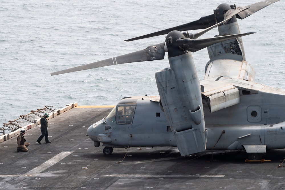 A Bell Boeing V-22 Osprey tiltrotor aircraft is parked on the flight deck of a naval vessel at sea. Two deck crew personnel are visible near the aircraft's nose, with one kneeling and one standing. The aircraft appears to be secured to the deck with tie-down chains, with rotors in a folded/transitional position.