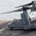 A Bell Boeing V-22 Osprey tiltrotor aircraft is parked on the flight deck of a naval vessel at sea. Two deck crew personnel are visible near the aircraft's nose, with one kneeling and one standing. The aircraft appears to be secured to the deck with tie-down chains, with rotors in a folded/transitional position.