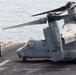 A Bell Boeing V-22 Osprey tiltrotor aircraft is parked on the flight deck of a naval vessel at sea. Two deck crew personnel are visible near the aircraft's nose, with one kneeling and one standing. The aircraft appears to be secured to the deck with tie-down chains, with rotors in a folded/transitional position.
