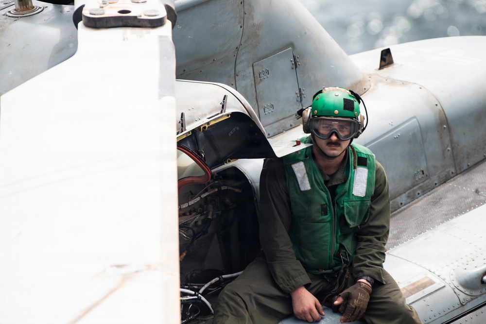 A military aviation deck crew member is seated on or near an aircraft, likely on the flight deck of a naval vessel. He is wearing standard naval aviation deck crew gear including a green helmet, goggles, green float coat/vest, and olive flight suit. Water is visible in the background, confirming a maritime/carrier deck environment.