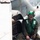 A military aviation deck crew member is seated on or near an aircraft, likely on the flight deck of a naval vessel. He is wearing standard naval aviation deck crew gear including a green helmet, goggles, green float coat/vest, and olive flight suit. Water is visible in the background, confirming a maritime/carrier deck environment.