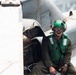 A military aviation deck crew member is seated on or near an aircraft, likely on the flight deck of a naval vessel. He is wearing standard naval aviation deck crew gear including a green helmet, goggles, green float coat/vest, and olive flight suit. Water is visible in the background, confirming a maritime/carrier deck environment.