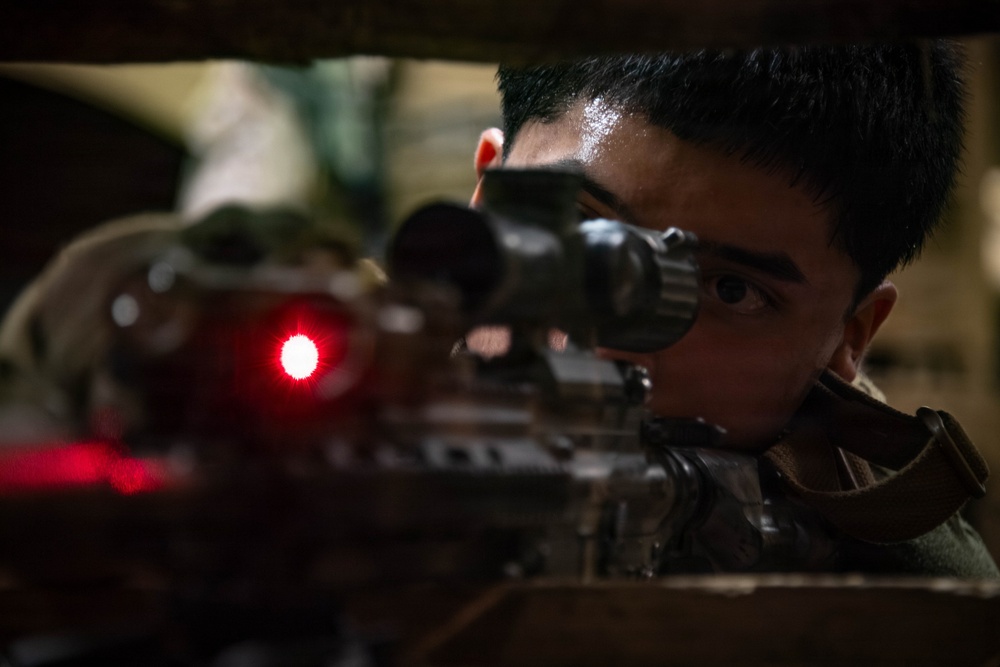 A young male in military or tactical gear is aiming down the scope of a rifle in a low-light environment. A red laser or illuminated dot is visible on the weapon, suggesting an active targeting or aiming device. The image is a close-up shot emphasizing the shooter's focused eye alignment with the optical scope.