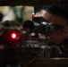 A young male in military or tactical gear is aiming down the scope of a rifle in a low-light environment. A red laser or illuminated dot is visible on the weapon, suggesting an active targeting or aiming device. The image is a close-up shot emphasizing the shooter's focused eye alignment with the optical scope.