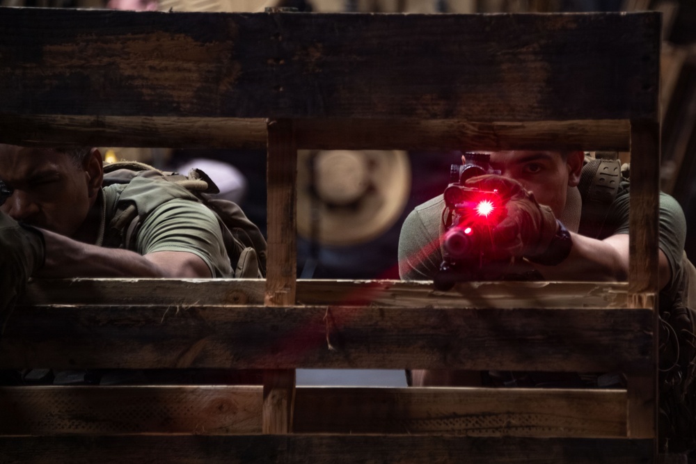 Two individuals in tactical/military-style clothing are positioned behind a wooden pallet or crate structure in a low-light environment. The person on the right is aiming a firearm equipped with a red laser sight directly toward the camera. The scene appears to be a training exercise, simulation, or film/media production based on the controlled indoor setting.