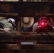 Two individuals in tactical/military-style clothing are positioned behind a wooden pallet or crate structure in a low-light environment. The person on the right is aiming a firearm equipped with a red laser sight directly toward the camera. The scene appears to be a training exercise, simulation, or film/media production based on the controlled indoor setting.