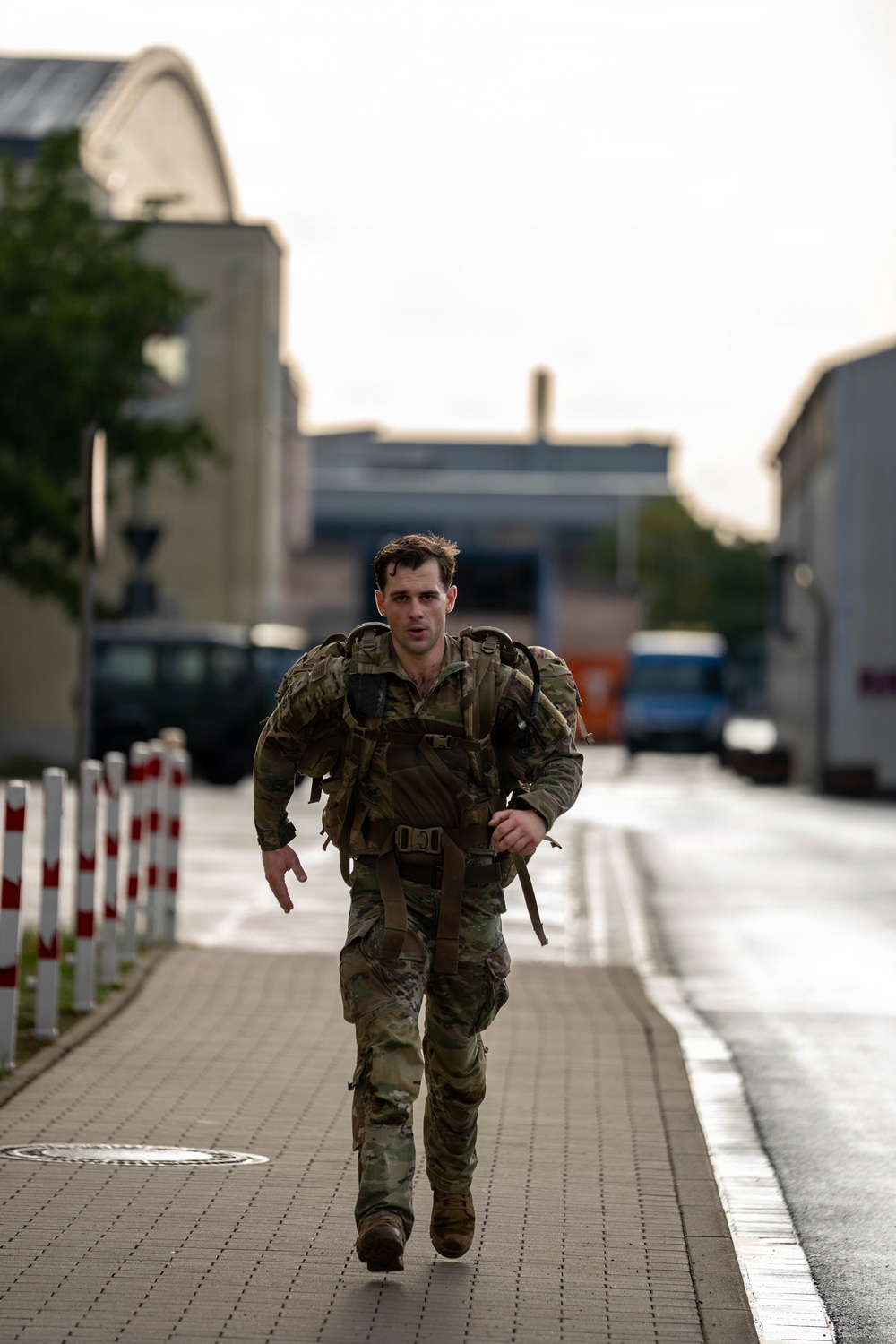 A male soldier in full camouflage uniform and tactical gear is running along a paved walkway in what appears to be a military installation or urban area. He is carrying a heavy rucksack and appears to be conducting a ruck march or physical training exercise. The wet pavement and overcast sky suggest recent rainfall.