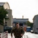 A male soldier in full camouflage uniform and tactical gear is running along a paved walkway in what appears to be a military installation or urban area. He is carrying a heavy rucksack and appears to be conducting a ruck march or physical training exercise. The wet pavement and overcast sky suggest recent rainfall.