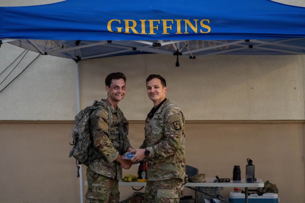 Two military personnel in U.S. Army OCP (Operational Camouflage Pattern) uniforms are posing together under a blue canopy tent labeled 'GRIFFINS' in gold lettering. One individual is wearing a backpack and they appear to be shaking hands or exchanging an item. The setting appears to be an outdoor military event or ceremony.