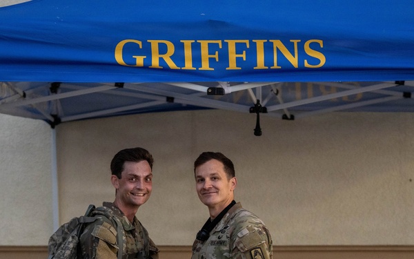 Two military personnel in U.S. Army OCP (Operational Camouflage Pattern) uniforms are posing together under a blue canopy tent labeled 'GRIFFINS' in gold lettering. One individual is wearing a backpack and they appear to be shaking hands or exchanging an item. The setting appears to be an outdoor military event or ceremony.