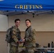 Two military personnel in U.S. Army OCP (Operational Camouflage Pattern) uniforms are posing together under a blue canopy tent labeled 'GRIFFINS' in gold lettering. One individual is wearing a backpack and they appear to be shaking hands or exchanging an item. The setting appears to be an outdoor military event or ceremony.