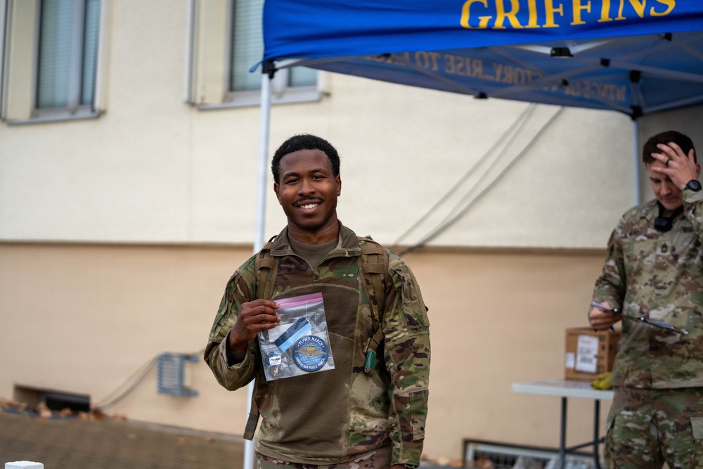A smiling military service member in camouflage uniform holds up a clear plastic bag containing what appears to be a patch and medal or ribbon. The event appears to be a 'Flyer March' associated with the US Air Force, indicated by the item in the bag and the blue canopy tent labeled 'GRIFFINS' in the background. A second service member is visible in the background near a table.