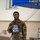 12th Combat Aviation Brigade hosts Danish Flyver March A smiling military service member in camouflage uniform holds up a clear plastic bag containing what appears to be a patch and medal or ribbon. The event appears to be a 'Flyer March' associated with the US Air Force, indicated by the item in the bag and the blue canopy tent labeled 'GRIFFINS' in the background. A second service member is visible in the background near a table.