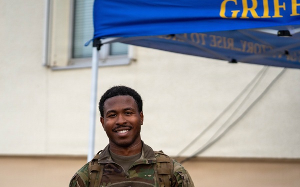 A smiling military service member in camouflage uniform holds up a clear plastic bag containing what appears to be a patch and medal or ribbon. The event appears to be a 'Flyer March' associated with the US Air Force, indicated by the item in the bag and the blue canopy tent labeled 'GRIFFINS' in the background. A second service member is visible in the background near a table.