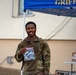 A smiling military service member in camouflage uniform holds up a clear plastic bag containing what appears to be a patch and medal or ribbon. The event appears to be a 'Flyer March' associated with the US Air Force, indicated by the item in the bag and the blue canopy tent labeled 'GRIFFINS' in the background. A second service member is visible in the background near a table.