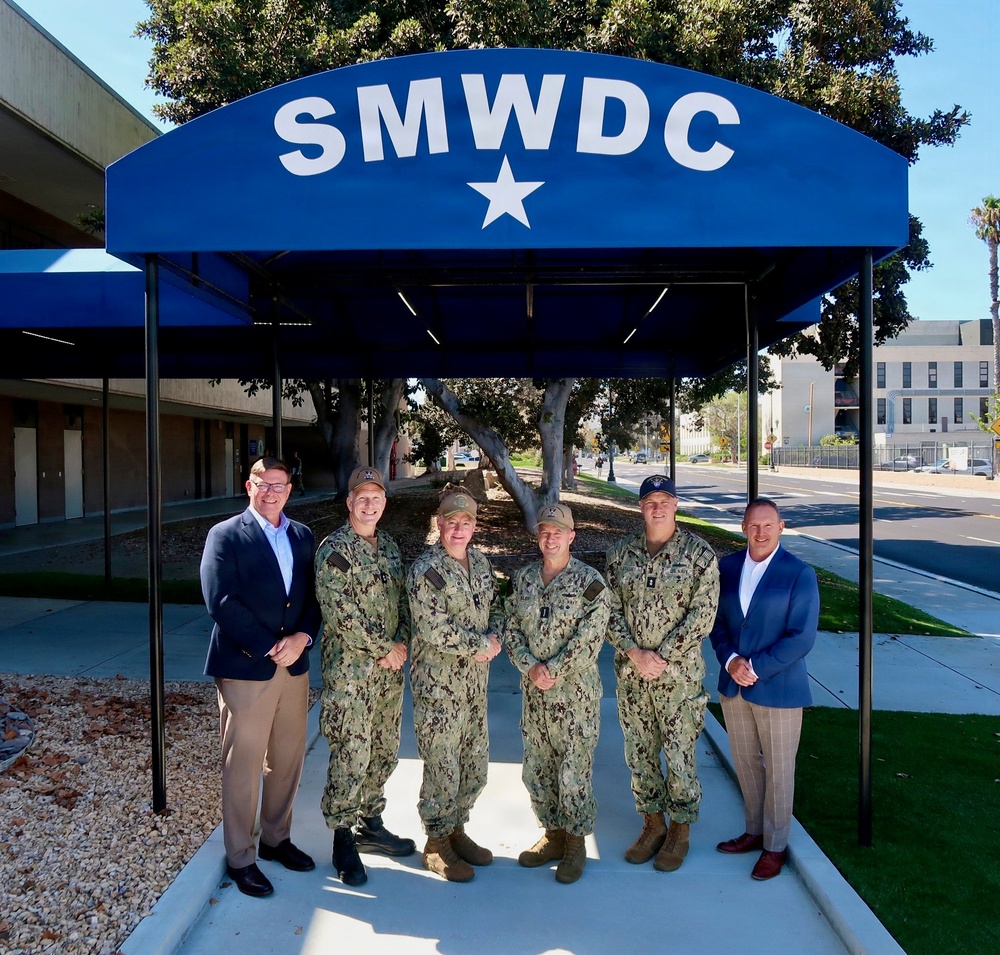 Six individuals pose for a group photo under a blue canopy bearing the acronym 'SMWDC' with a single white star. Four individuals are wearing U.S. Navy camouflage uniforms (NWU Type III), while two individuals on the far left and far right are dressed in civilian business attire. The setting appears to be an outdoor entrance area of a military facility, likely Naval Base San Diego.