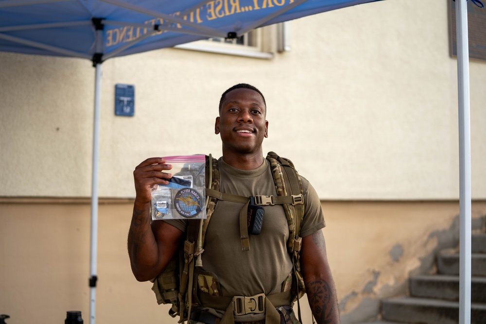 A smiling military service member wearing an olive drab t-shirt and tactical gear with a large backpack stands under a blue canopy tent. He is holding up a clear plastic zip-lock bag containing what appears to be patches and memorabilia, one of which reads 'FLYER MARCH' and appears to reference the Air Force. The setting appears to be an outdoor military event or competition.