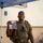 12th Combat Aviation Brigade hosts Danish Flyver March A smiling military service member wearing an olive drab t-shirt and tactical gear with a large backpack stands under a blue canopy tent. He is holding up a clear plastic zip-lock bag containing what appears to be patches and memorabilia, one of which reads 'FLYER MARCH' and appears to reference the Air Force. The setting appears to be an outdoor military event or competition.