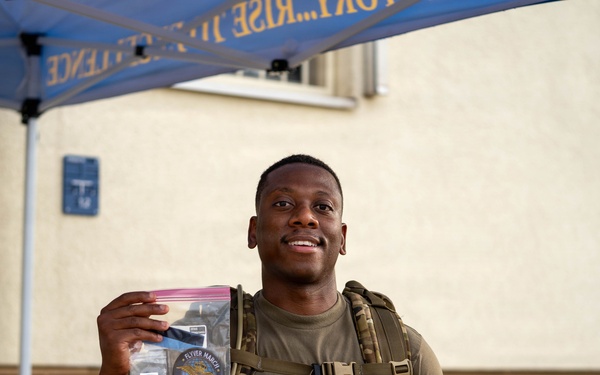 A smiling military service member wearing an olive drab t-shirt and tactical gear with a large backpack stands under a blue canopy tent. He is holding up a clear plastic zip-lock bag containing what appears to be patches and memorabilia, one of which reads 'FLYER MARCH' and appears to reference the Air Force. The setting appears to be an outdoor military event or competition.