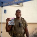 A smiling military service member wearing an olive drab t-shirt and tactical gear with a large backpack stands under a blue canopy tent. He is holding up a clear plastic zip-lock bag containing what appears to be patches and memorabilia, one of which reads 'FLYER MARCH' and appears to reference the Air Force. The setting appears to be an outdoor military event or competition.