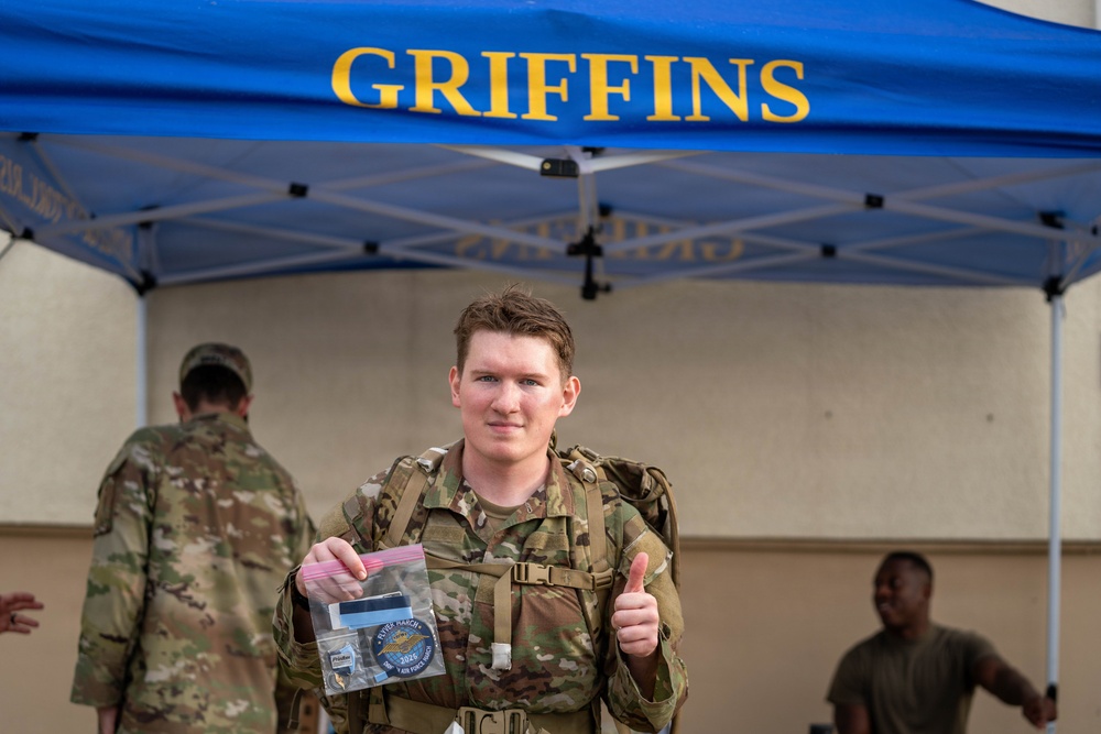 A military service member in OCP (Operational Camouflage Pattern) uniform gives a thumbs-up while holding a clear plastic bag containing what appears to be a patch or commemorative item from a ruck march event. They are standing in front of a blue canopy tent labeled 'GRIFFINS,' likely associated with a U.S. Air Force unit. The setting appears to be an outdoor military event or competition, possibly a ruck march completion ceremony.