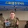 12th Combat Aviation Brigade hosts Danish Flyver March A military service member in OCP (Operational Camouflage Pattern) uniform gives a thumbs-up while holding a clear plastic bag containing what appears to be a patch or commemorative item from a ruck march event. They are standing in front of a blue canopy tent labeled 'GRIFFINS,' likely associated with a U.S. Air Force unit. The setting appears to be an outdoor military event or competition, possibly a ruck march completion ceremony.