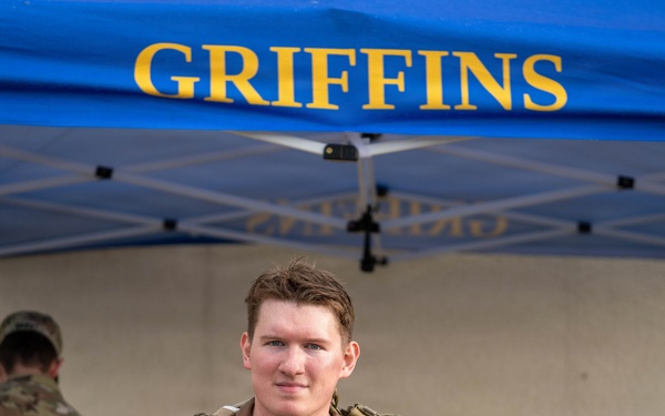 A military service member in OCP (Operational Camouflage Pattern) uniform gives a thumbs-up while holding a clear plastic bag containing what appears to be a patch or commemorative item from a ruck march event. They are standing in front of a blue canopy tent labeled 'GRIFFINS,' likely associated with a U.S. Air Force unit. The setting appears to be an outdoor military event or competition, possibly a ruck march completion ceremony.