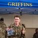 A military service member in OCP (Operational Camouflage Pattern) uniform gives a thumbs-up while holding a clear plastic bag containing what appears to be a patch or commemorative item from a ruck march event. They are standing in front of a blue canopy tent labeled 'GRIFFINS,' likely associated with a U.S. Air Force unit. The setting appears to be an outdoor military event or competition, possibly a ruck march completion ceremony.