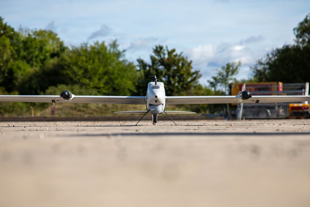 A fixed-wing UAV (unmanned aerial vehicle) is positioned on a paved surface, photographed from a low front-facing angle. The drone features a pusher propeller configuration at the rear, two wing-mounted sensor pods, and a tricycle-style landing gear. The background shows green trees, a partly cloudy sky, and what appears to be a fenced industrial or airfield area.