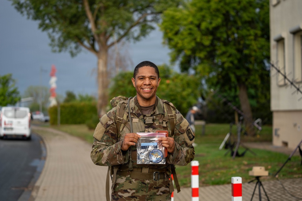 A smiling U.S. Army soldier in MultiCam/OCP camouflage uniform is posing outdoors while holding a clear plastic bag containing what appears to be military patches or insignia. He is wearing a rucksack and standing on a brick pathway near a building, trees, and red-and-white bollards. The setting appears to be a military installation or base, likely in Europe.